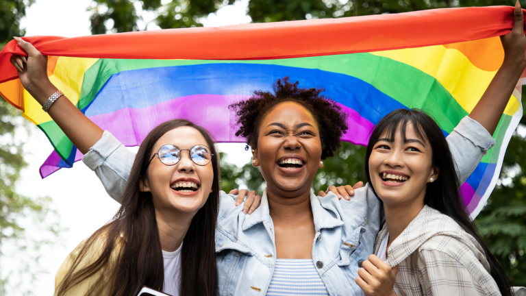 Three multicultural Gen Z women under a pride flag